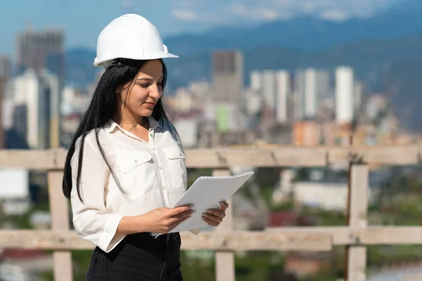 stock image Young female architect in a hard hat on a construction site while working with a paper tablet. Supervisor wearing a protective helmet while working on a construction site. Successful inspector.