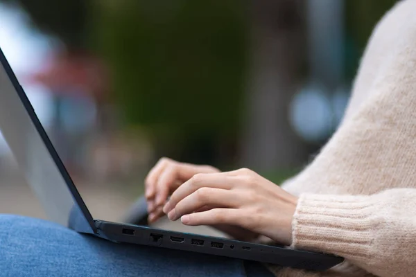 Close-up of female hands of a professional user-an employee typing on a laptop, sitting on a park bench. Student, freelancer at work with a laptop in the park