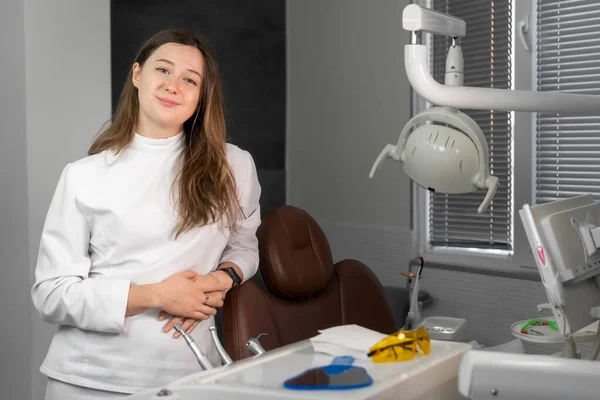 Beyaz takım elbiseli genç, gülümseyen kadın dişçi, dişçi koltuğunun yanında duruyor ve kameraya bakıyor.