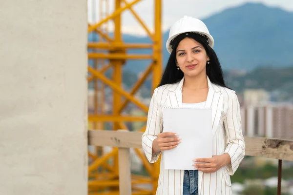 年轻美丽的 面带微笑的高加索女建筑师 头戴防护头盔 身穿夹克 站在建筑工地上 用的是纸片 — 图库照片
