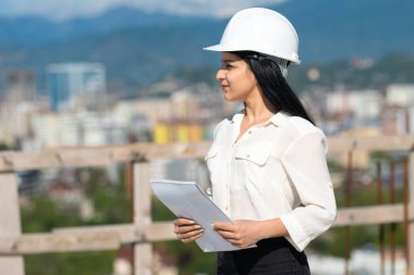 Bir inşaat alanında kağıt tabletle çalışırken kasketli genç bir kadın mimar başka tarafa bakıyor. İnşaat alanında çalışırken koruyucu kask takan bir müfettiş..