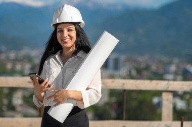 Güzel, genç, gülümseyen bir mimar, beyaz bir mimar, cep telefonu ve ellerinde bir çizimle dağların arka planında bir inşaat alanında. İnşaat konsepti