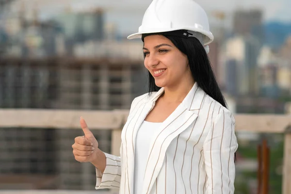 Young beautiful smiling woman in a white helmet shows a thumbs up sign, OK on a construction site. Concept of the profession of architect, engineer, manager, surveyor
