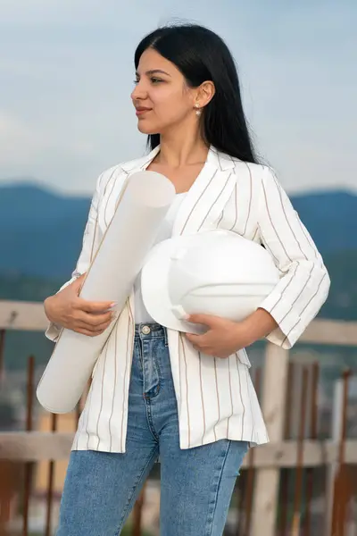 Young beautiful woman, an engineer, manager or architect, holds a white helmet for safe work and a drawing pad with drawings on the construction site. Concept of building,development,construction