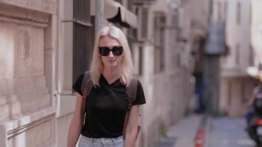 Sırt çantalı, siyah güneş gözlüğü takan sarışın bir kadının ön görüntüsü. Yürüyor, eski boş caddede yürüyor ve seyahatin ve yeni yerin tadını çıkarıyor, gezinin tadını çıkarıyor. Yol boyunca yeşil alanlar ve ağaçlar.