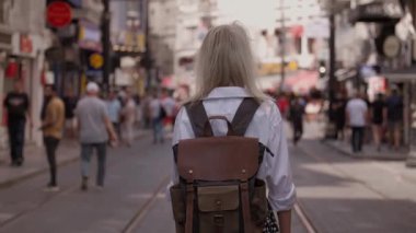 Sırt çantası ve beyaz gömleğiyle yaz modası tarzında sokakta yürüyen güzel sarışın kadın turistin arka görüntüsü. Yeni şehri keşfeden çekici bir kız. Sunny Image Moda Portresi