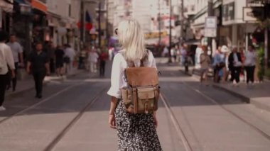 Sırt çantası ve beyaz gömleğiyle yaz modası tarzında sokakta yürüyen güzel sarışın kadın turistin arka görüntüsü. Yeni şehri keşfeden çekici bir kız. Sunny Image Moda Portresi