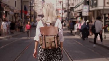 Genç, güzel, narin bir kadın işlek şehir caddesinde yürüyor. Şık seyahat kıyafetleri içinde turist, beyaz gömlek ve sırt çantasıyla gezer, yeni yerler ve sokaklar keşfeder..