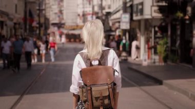 Sırt çantası ve beyaz gömleğiyle yaz modası tarzında sokakta yürüyen güzel sarışın kadın turistin arka görüntüsü. Yeni şehri keşfeden çekici bir kız. Sunny Image Moda Portresi