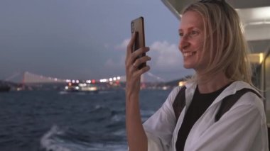 Akşamları ya da gece feribotla seyahat ederken deniz ve kıyı kentinin fotoğraflarını çeken genç bir kadın. Şehrin ışıkları yanıyordu. Tekne gezisi sırasında akıllı telefonunda kamera kullanmak.