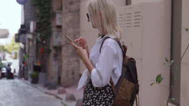 Sırt çantalı turist kadın şehir caddesinde akıllı telefon kullanıyor. Genç bayan gezgin akıllı telefon kullanarak sokaklarda yürüyor. Koyu gözlüklü sarışın ve sırt çantalı bağımsız yürüyüşler yapıyor.