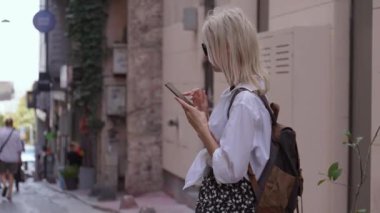 Sırt çantalı turist kadın şehir caddesinde akıllı telefon kullanıyor. Genç bayan gezgin akıllı telefon kullanarak şehrin sokaklarında yürüyor. Güneş gözlüklü sarışın kadın ve sırt çantalı taksi çağırma uygulaması.