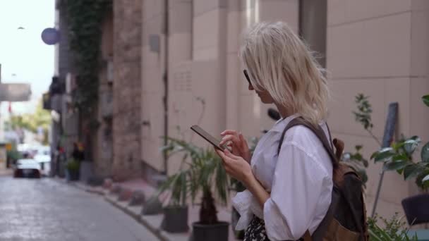 スタイリッシュな服とバックパック付きの観光客は ホテルのサインの背景に対してストリートに立っており 地図上のアドレス位置のためのアプリケーションを探している彼女の携帯電話を見ます 宿泊施設 ホテル ファインダー — ストック動画
