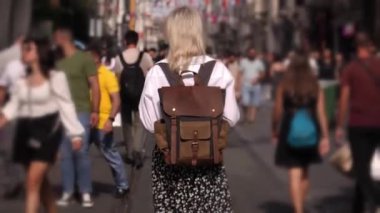 Genç, güzel, narin bir kadın işlek şehir caddesinde yürüyor. Şık seyahat kıyafetleri içinde turist, beyaz gömlek ve sırt çantasıyla gezer, yeni yerler ve sokaklar keşfeder..
