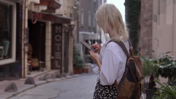 スタイリッシュな服とバックパック付きの観光客は ホテルのサインの背景に対してストリートに立っており 地図上のアドレス位置のためのアプリケーションを探している彼女の携帯電話を見ます 宿泊施設 ホテル ファインダー — ストック動画