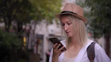 Beyaz tenli, şapka ve gömlekli, akıllı telefona bakan, şehirde yürürken gülümseyen ve tatilin tadını çıkaran turist bir kadın. Şehir konumu olan taksi haritaları uygulamasında arama yapar