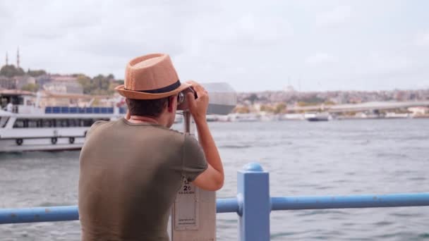 コインで双眼鏡を通して都市を眺める帽子とバックパックの若い観光客 ボスポラスの船やボートを通過するイスタンブールの望遠鏡を通って探している男または男 — ストック動画