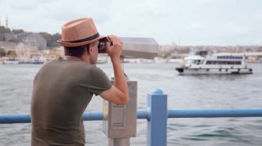 Seyahat: Şapkalı ve sırt çantalı genç turist madeni paralı dürbünle şehre bakıyor. İstanbul 'da teleskopla Boğaz' dan geçen gemi ve teknelere bakan adam ya da adam..