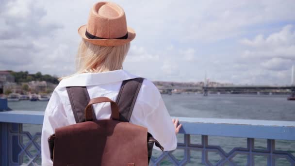 土耳其伊斯坦布尔的一座桥上站着一位漂亮的年轻女子 她背着背包和草帽 看着船只在博斯普鲁斯海峡上航行 探索城市和旅行 — 图库视频影像