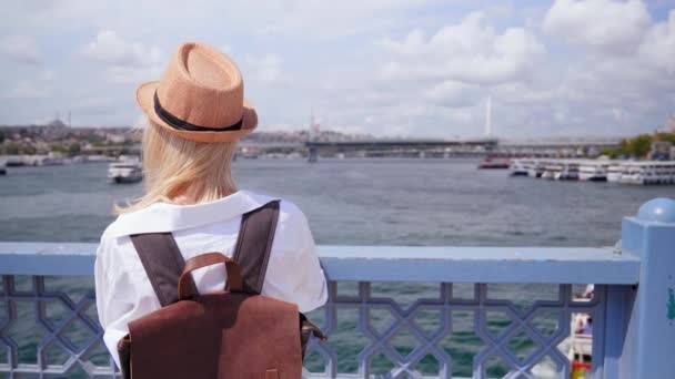 土耳其伊斯坦布尔的一座桥上站着一位漂亮的年轻女子 她背着背包和草帽 看着船只在博斯普鲁斯海峡上航行 探索城市和旅行 — 图库视频影像