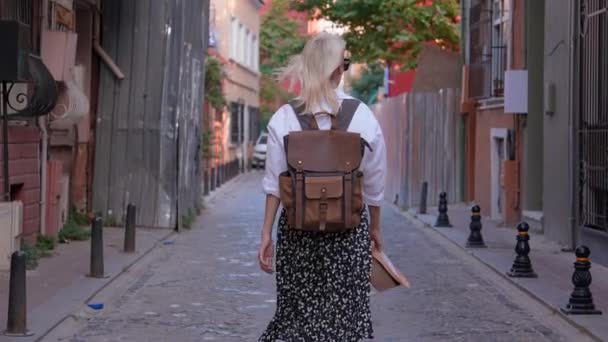 穿着白衬衫 背着背包 头戴破帽子的金发女游客的背影 她走在空荡荡的街道上 享受着旅行和观光的乐趣 旧城的小街 — 图库视频影像