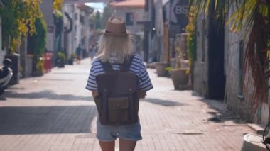 Beyaz tişörtlü, sırt çantası ve kırık şapkalı sarışın bir kadın. Boş caddede yürüyor ve gezmekten, yeni bir yerden ve gezmekten hoşlanıyor. Eski kasabadaki küçük işlek cadde.