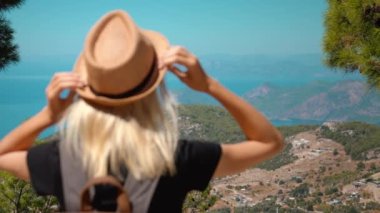 Sırt çantalı ve şapkalı kız dağın tepesinden deniz, okyanus ve adaları olan güzel dağ vadisine bakıyor. Kayanın üzerinde oturan genç, sarışın bir kadınla manzara. Yürüyüş. Doğa