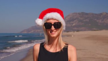 Noel 'de Noel Baba şapkalı genç bir kadının yakın çekimi ya da yeni yılda sahilde güneş gözlüklü dağların arka planında. Dağları gören bir kız kıyıda yürüyor. Ziyafete Yolculuk.