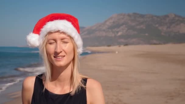 サンタクロースの帽子をかぶった若い白人女性のクローズアップ クリスマスやバックグラウンドの山と海岸の海岸で新年 山を眺めながら沿岸を歩いている少女 トリップと休日 — ストック動画