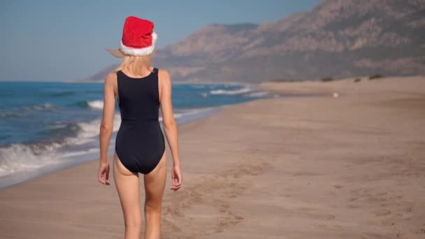 サンタクロースの帽子の若い白人女性のバックビュー クリスマスやバックグラウンドの山と海岸の海岸で新年 山を眺めながら沿岸を歩いている少女 トリップと休日 — ストック動画