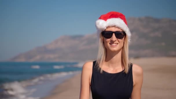 圣诞或新年的时候 年轻女子戴着圣爪帽在海滨的特写镜头 戴着太阳镜背景的山 女孩走在海岸上俯瞰群山 佳肴之旅 — 图库视频影像