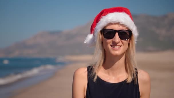 サンタクロースの帽子の若い女性のクローズアップ クリスマスやサングラスの山の背景の海岸の海岸で新年 山を眺めながら沿岸を歩いている少女 フィーストへの旅 — ストック動画