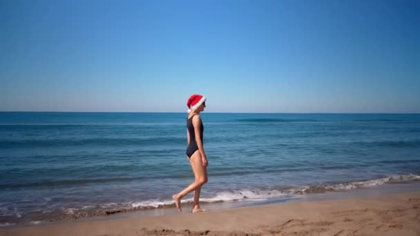 サンタクロースの帽子をかぶった若い女性のクローズアップは サングラスの海の背景に対して海岸の海岸でクリスマスや新年に行われます 海を眺めながら海岸を歩いている少女 ホリデー — ストック動画