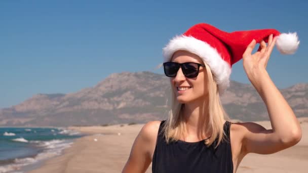 サンタクロースの帽子をかぶった若い女性のクローズアップは サングラスの海の背景に対して海岸の海岸でクリスマスや新年に行われます 彼女の帽子に触れる山の景色と沿岸を歩いている少女 — ストック動画