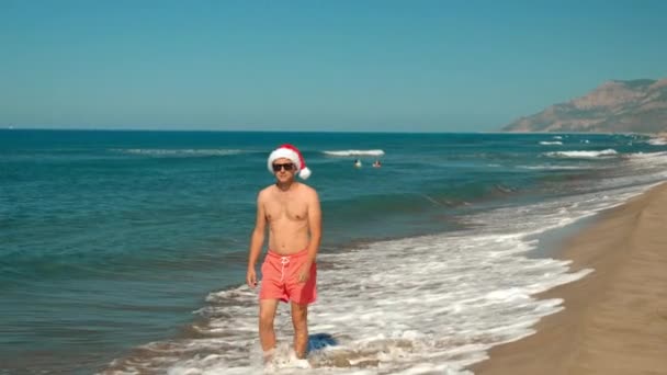 男人的特写镜头 在圣诞或新年的时候戴圣爪帽的家伙在海滨与海浪在山上背景的太阳镜 沿着海岸走 可以看到群山 度假之旅 — 图库视频影像