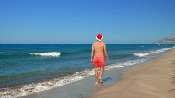 圣诞节或新年的时候 年轻女人或男人戴着圣爪帽在海滨的背景图片 沿着海岸走 可以看到群山 旅行和休息 炎热温暖的地方 — 图库视频影像