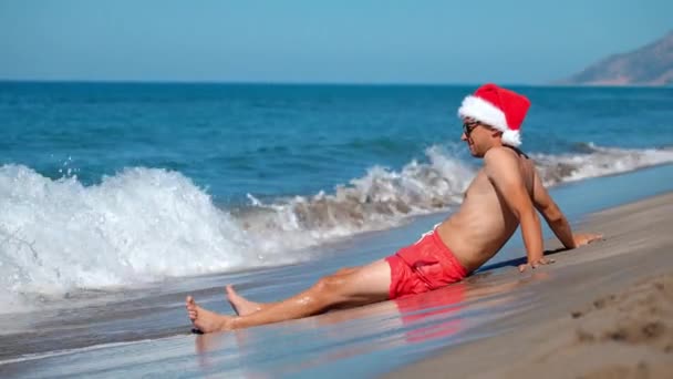 サングラスの若いハンサムな男は 晴れた日サンタビーチでサンタクロースの帽子を浴びています 新しいおめでとうございます ホリデーリゾート冬 12月のホットな国や場所でのクリスマス 山の波 — ストック動画