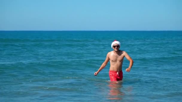 サンタクロースマン リトルレッドライディングフードランニングで ダンスと馬鹿げた周り 彼のフィートとスプラッシュで水を投げる ビーチハッピーサングラス バックグラウンド海岸の山々に対して クリスマス — ストック動画
