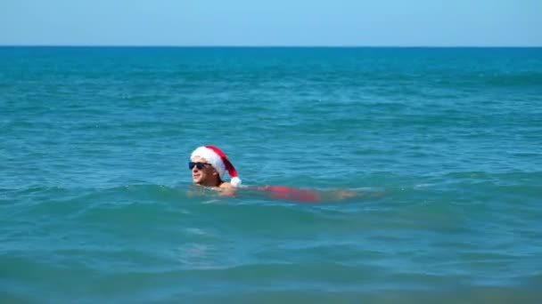 サンタクロースの帽子をかぶった男 サングラスの波で 海岸に座って 彼はクリスマスか新年の日に海で泳いでいます トラベルホリデー 12月の温水リゾートでの入浴 — ストック動画