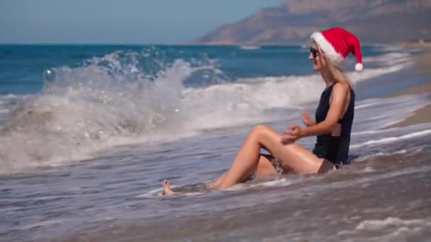 砂浜でサンタクロースの帽子で日光浴で美しいスレンダーの女性は 晴れた日を楽しんでいます 新しいおめでとうございます ホットな国や場所で12月のクリスマスリゾート — ストック動画