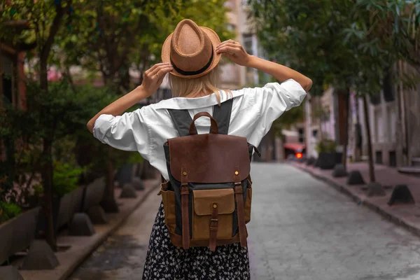 Beyaz gömlekli şık bir genç kadın, sırt çantalı bir etek, Türkiye 'de güzel bir caddenin ortasında dururken şapkasını elleriyle tutuyor. Seyahat kavramı, turizm. Dikey fotoğraf