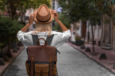 Beyaz gömlekli şık bir genç kadın, sırt çantalı bir etek, Türkiye 'de güzel bir caddenin ortasında dururken şapkasını elleriyle tutuyor. Seyahat kavramı, turizm. Dikey fotoğraf