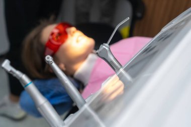 Önleyici muayene için dişçi randevusunda sandalyede oturan genç ve güzel bir hastanın bulanık arka planında diş aletlerinin yakın plan görüntüsü.