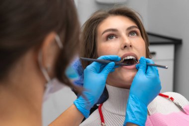 Kadın dişçi, bir diş kliniğinde aletlerin yardımıyla hastanın ağız boşluğunu muayene eder. Genç hasta, dişçi koltuğunda ağzı açık bir şekilde muayene için oturuyor..