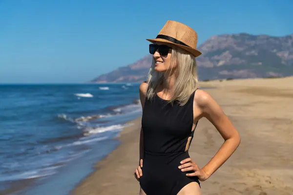 Smiling blonde woman in a black one-piece swimsuit, hat, sunglasses standing on the beach of the sea. Young smiling woman on vacation enjoys the sea breeze. Attractive slender beautiful girl.