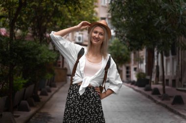 Etek, gömlek, şapka ve sırt çantasıyla güzel bir sokakta poz veren güzel sarışın turist kadın kameraya bakar. Seyahat, tatil, Türkiye.
