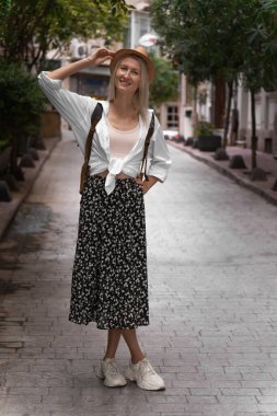 Etek, gömlek, şapka ve sırt çantasıyla güzel bir sokakta poz veren güzel sarışın turist kadın kameraya bakar. Seyahat, tatil, Türkiye kavramı. Dikey fotoğraf