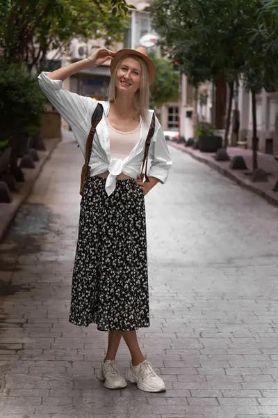 Etek, gömlek, şapka ve sırt çantasıyla güzel bir sokakta poz veren güzel sarışın turist kadın kameraya bakar. Seyahat, tatil, Türkiye kavramı. Dikey fotoğraf