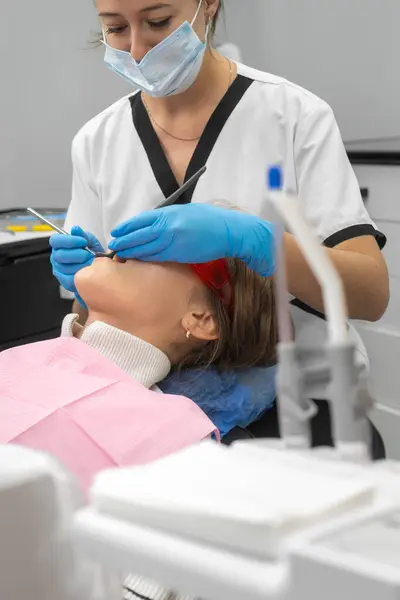 Female dentist in a uniform, medical mask examines the teeth of a patient in a dental clinic. Attractive patient lying on a dental chair receives dental treatment from a dentist. Vertical photo