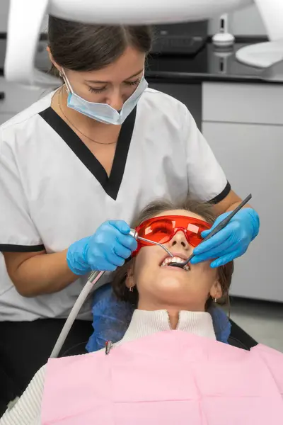 Woman dentist washes the patient\'s teeth during an oral examination. Young woman wearing safety glasses in a dental chair at a dentist appointment. Vertical photo